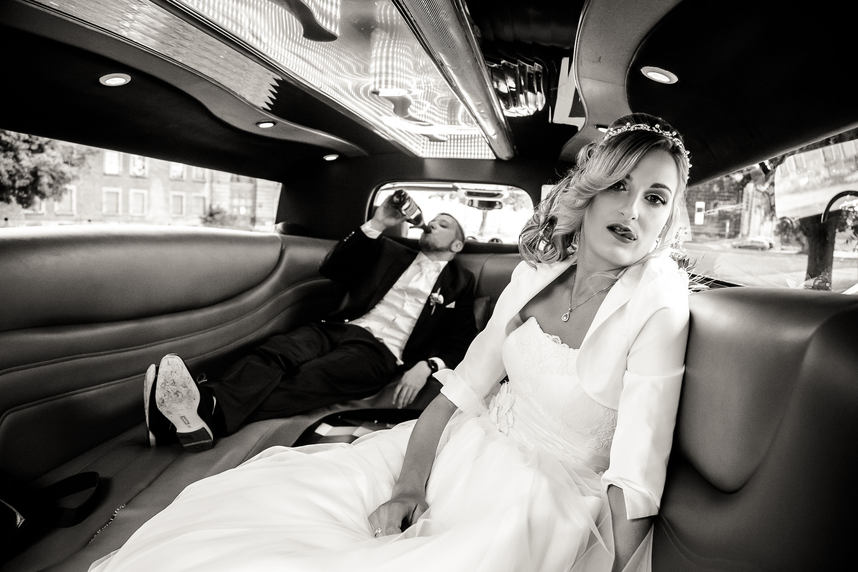 Sie hat "Ja" gesagt, er hat Durst: die Braut im Hochzeitskleid posiert schick in der Limo, während der Bräutigam im Anzug hinten lässig aus der Flasche Champus trinkt. Prioritäten im Hochzeitsauto klar verteilt – Glamour vorne, Chillen (mit Getränk) hinten. Canon 6d & EF 24-105 f4.0