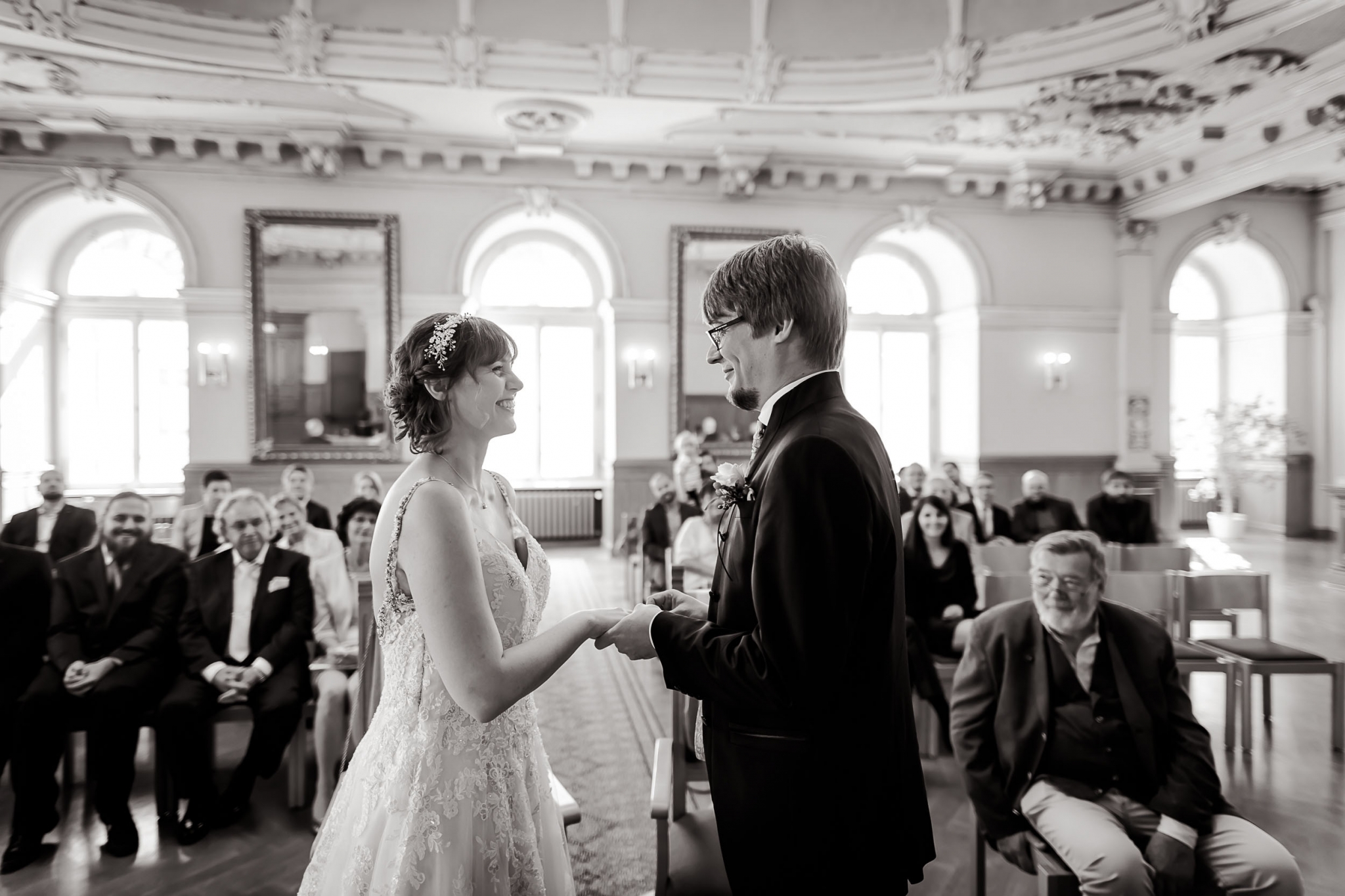 Ein Augenblick inniger Verbundenheit: das Hochzeitspaar hält sich liebevoll die Hände und tauscht verliebte Blicke und die Ringe in einem feierlichen Moment. Umgeben von den Zeugen ihrer Liebe, versprechen sie sich eine gemeinsame Zukunft voller Romantik und Glück