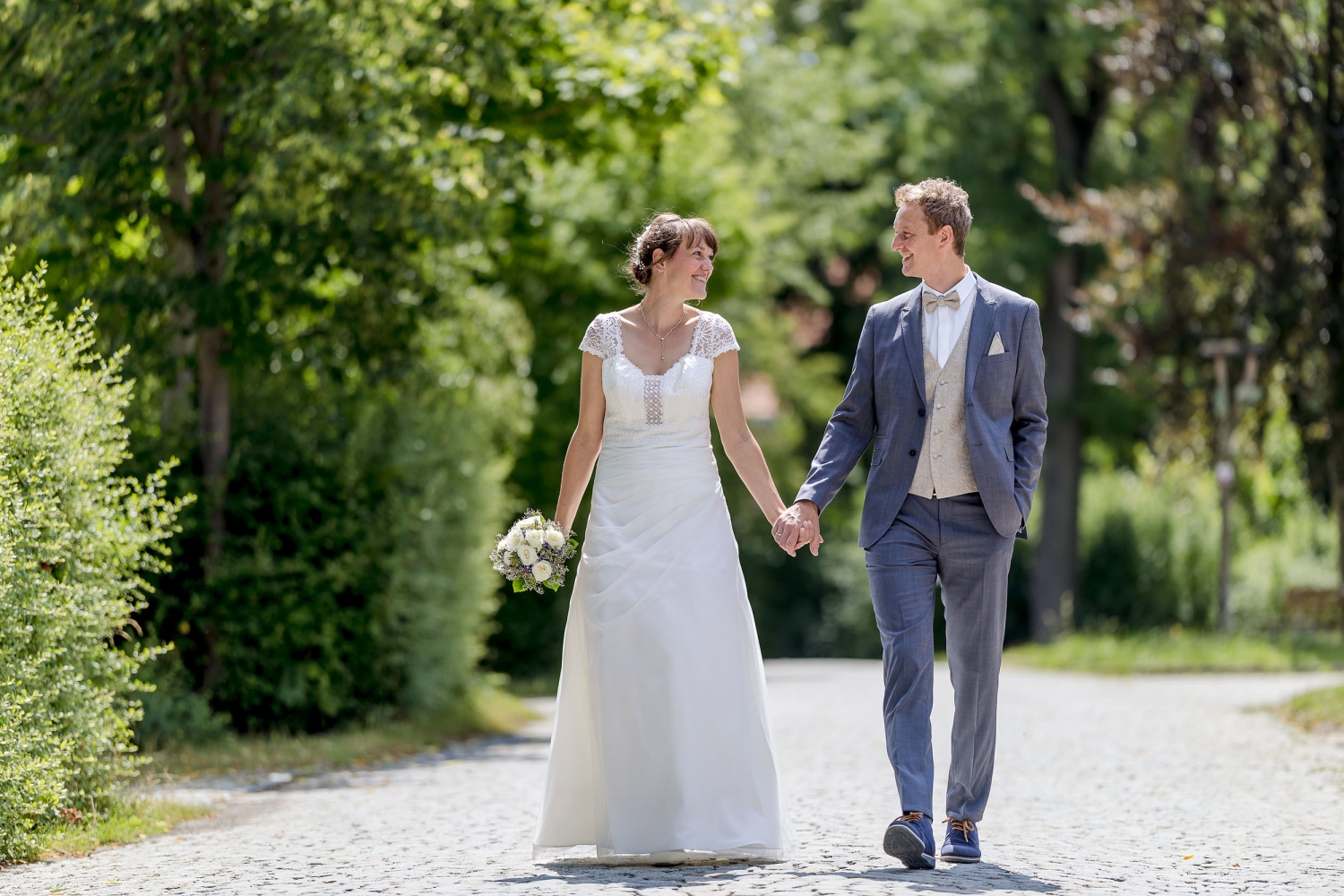 Ein glückliches Brautpaar spaziert Hand in Hand auf einem von Bäumen gesäumten Weg ins gemeinsame Glück, fotografiert in Kranichfeld bei Erfurt mit der EOS R5M² & dem RF 135mm f1.8