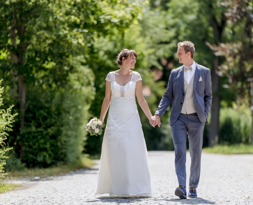 Ein glückliches Brautpaar spaziert Hand in Hand auf einem von Bäumen gesäumten Weg ins gemeinsame Glück, fotografiert in Kranichfeld bei Erfurt mit der EOS R5M² & dem RF 135mm f1.8