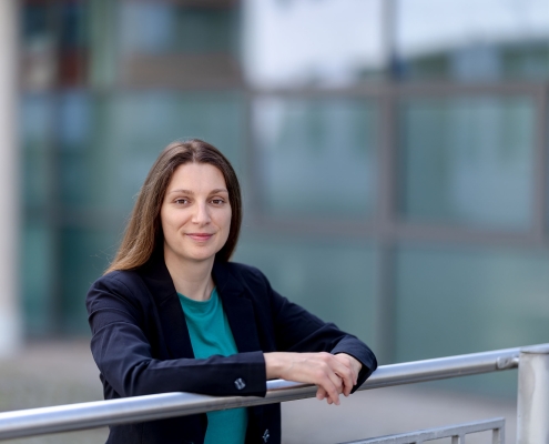 Businessportrait einer Frau mit braunen Haaren und einem freundlichen Blick. Sie steht in einem städtischen Umfeld vor einer verschwommenen Glasfassade fotografiert in Erfurt mit der Canon EOS R5m2 & dem RF 85mm f1.2