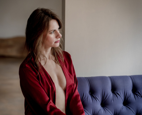 Sinnliches Boudoir-Porträt: Frau im roten Seidenmantel Sinnliches Boudoir-Porträt einer brünetten Frau, die in einem offenen, tiefroten Seidenmorgenmantel auf einem blauen Chesterfield-Samtsofa sitzt und verträumt aus dem Fenster blickt. Canon EOS R5M2 & RF85mm f1.2