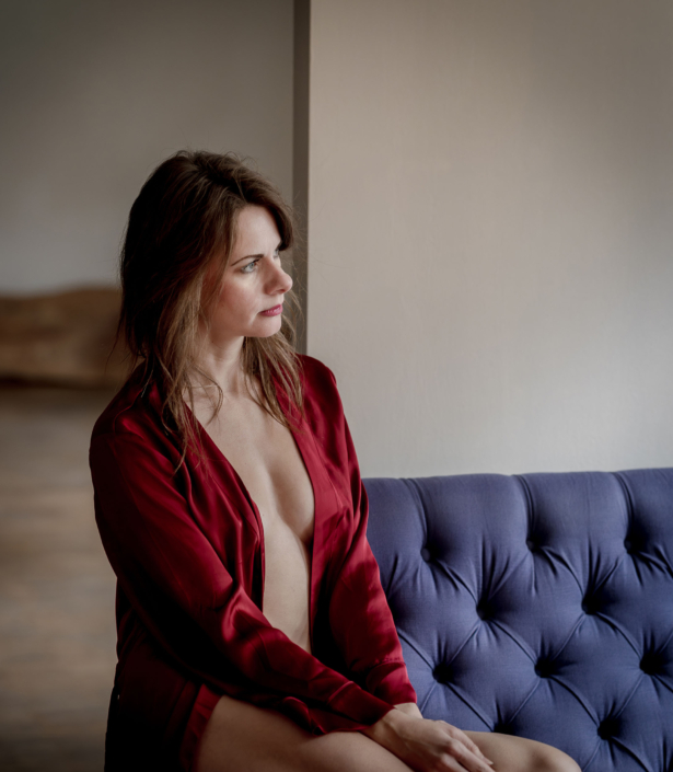 Sinnliches Boudoir-Porträt: Frau im roten Seidenmantel Sinnliches Boudoir-Porträt einer brünetten Frau, die in einem offenen, tiefroten Seidenmorgenmantel auf einem blauen Chesterfield-Samtsofa sitzt und verträumt aus dem Fenster blickt. Canon EOS R5M2 & RF85mm f1.2