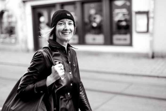 Lächelnde dynamische Frau im Urban Streetstyle Schwarzweiß Lächelnde Frau in schwarzer Lederjacke und Mütze auf einer Einkaufstraße in seitlicher Bewegung fotografiert