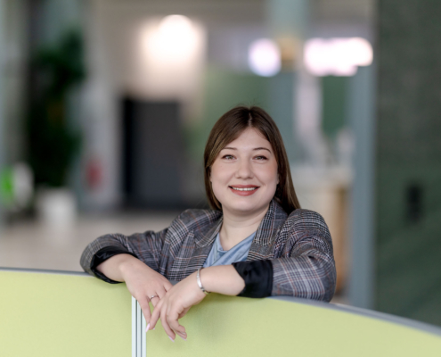 Business-Portraet-Mitarbeiterin-Buero Lächelnde Mitarbeiterin im karierten Blazer lehnt an einer Bürotrennwand.