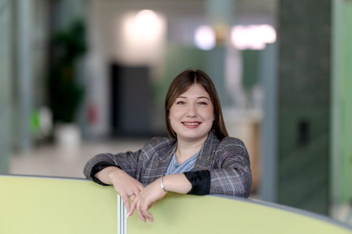 Business-Portraet-Mitarbeiterin-Buero Lächelnde Mitarbeiterin im karierten Blazer lehnt an einer Bürotrennwand.