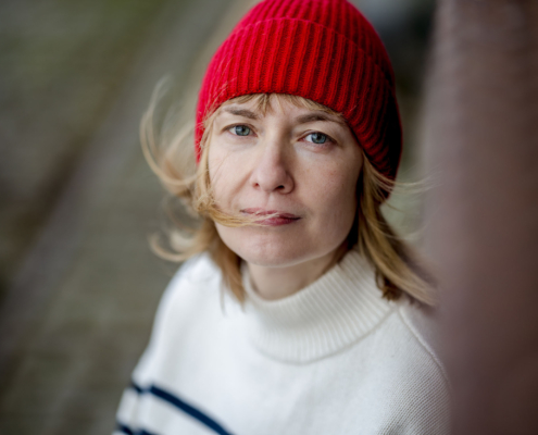 Authentisches Frauenporträt an einem windigen Tag mit einer roten Mütze als Farbakzent. Die Aufnahme fängt einen nachdenklichen Moment in urbaner Umgebung ein.