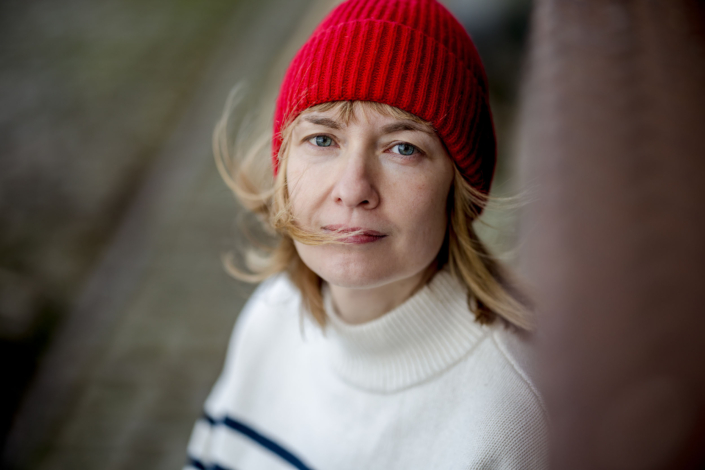 Authentisches Porträt einer Frau mit roter Beanie Authentisches Frauenporträt an einem windigen Tag mit einer roten Mütze als Farbakzent. Die Aufnahme fängt einen nachdenklichen Moment in urbaner Umgebung ein.