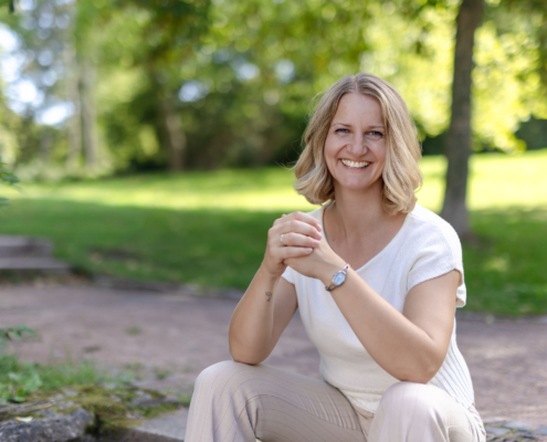 Eine blonde Frau in heller Kleidung schaut im Grünen lächelnd in die Kamera