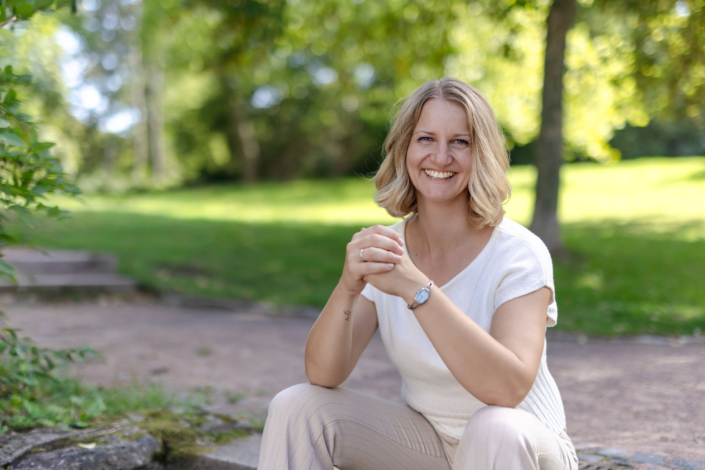 Eine blonde Frau in heller Kleidung schaut im Grünen lächelnd in die Kamera